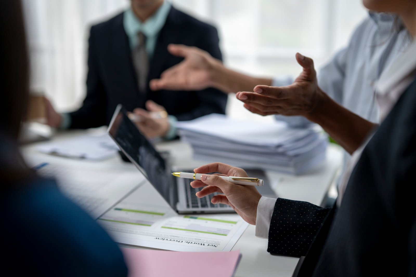 A group of people are sitting around a table with a laptop and a pen